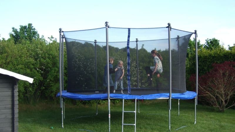Three kids joyfully bounce on a safe trampoline with a safety net, nestled in a backyard surrounded by trees.