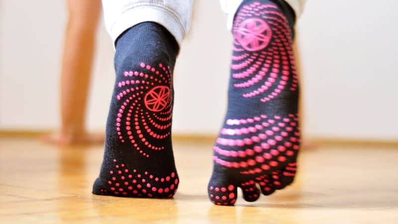 A person in black socks with pink dots stands on a wooden floor. To wash grip socks hassle-free and keep them vibrant, follow these steps.
