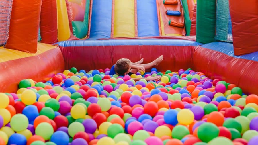 A child joyfully plays in a colorful ball pit at a trampoline park, featuring inflatable walls, a slide, and safe fun.